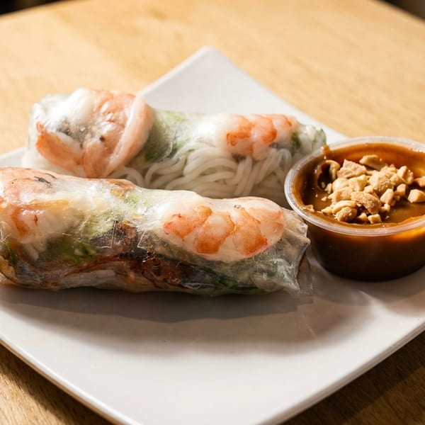 Two fresh shrimp and noodle spring rolls with a side of peanut sauce on a white plate.